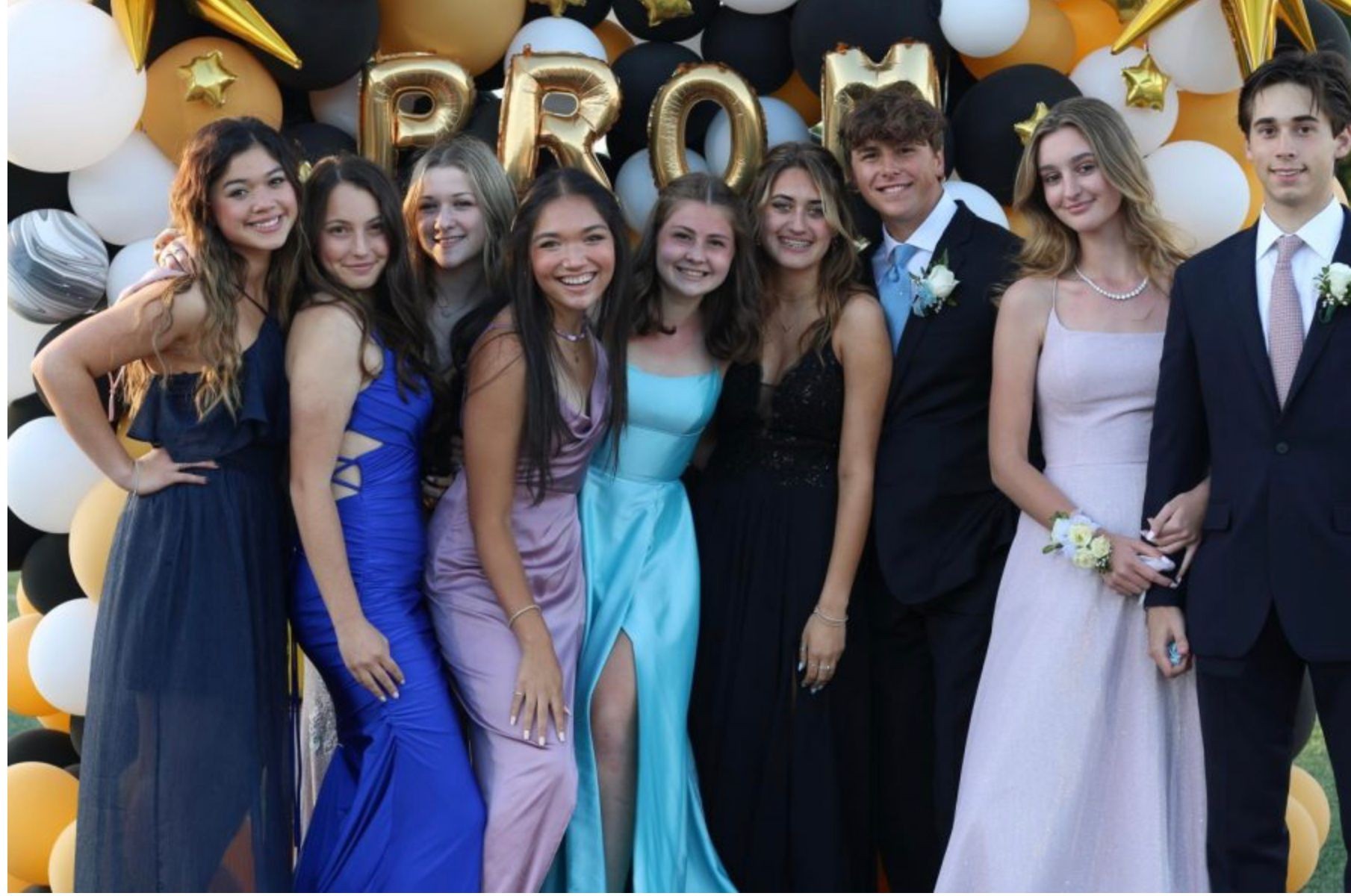 Group of people in formal attire posing in front of a balloon arch with gold letters spelling PROM.